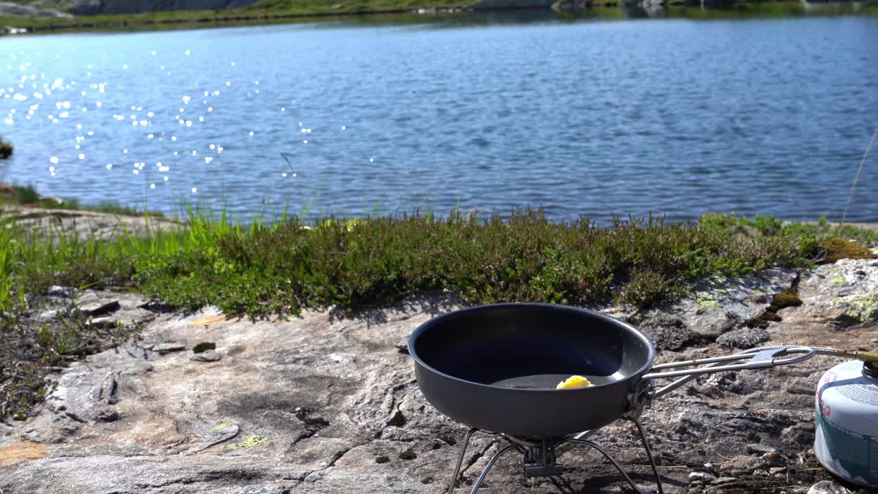 Person lights camping stove to melt butter in skillet, lake in background