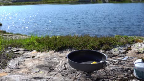 person lights camping stove to melt butter in skillet, lake in background