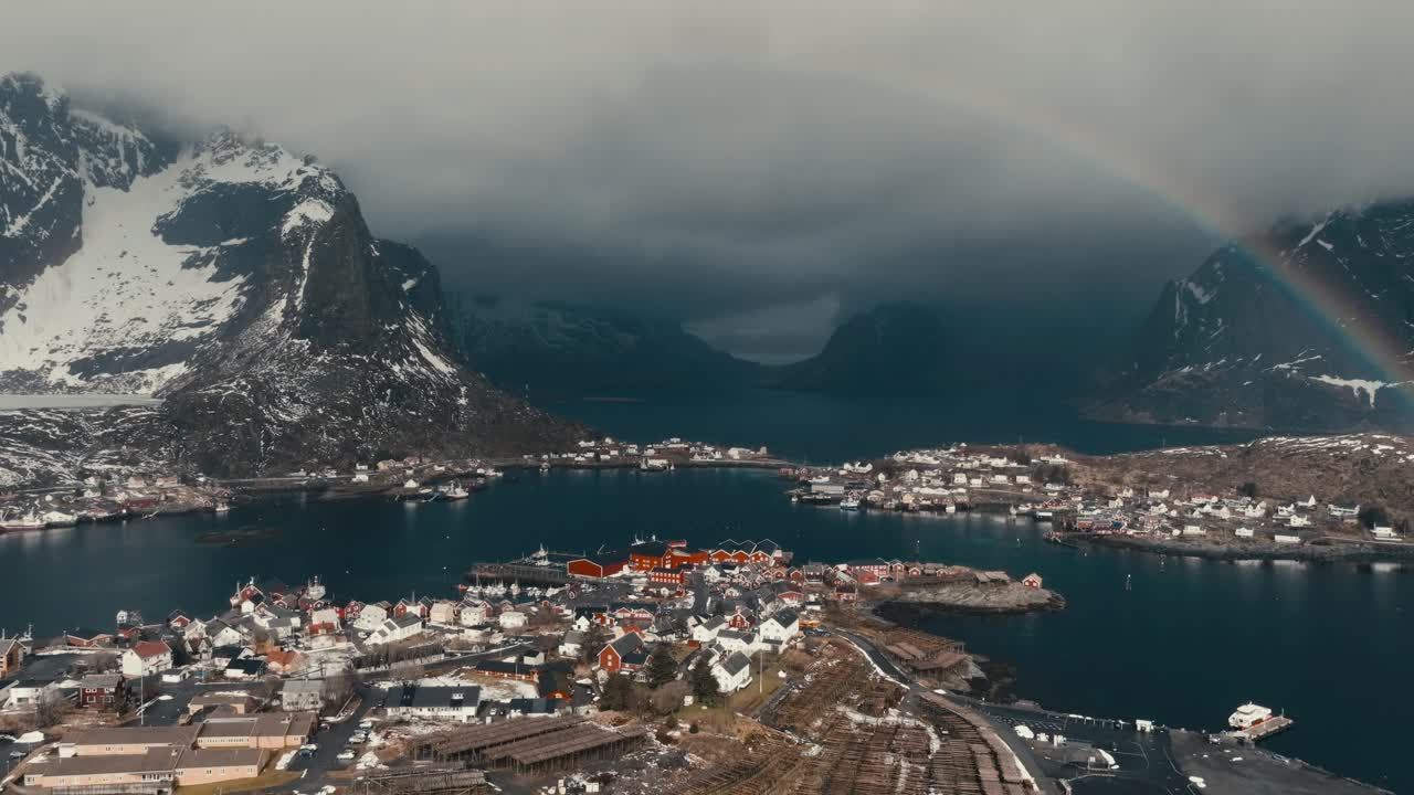 Fishing Village In Lofoten Islands Surrounded By Snowy Mountains Under Cloudy Sky With Rainbow. Reine, Norway. aerial pullback shot