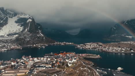 fishing village in lofoten islands surrounded by snowy mountains under cloudy sky with rainbow. reine, norway. aerial pullback shot
