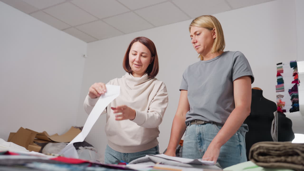 Clothes designers lifting cut out pattern piece during brainstorming session, carefully examining design shape and structure while surrounded by sewing tools, materials, and mirror in background
