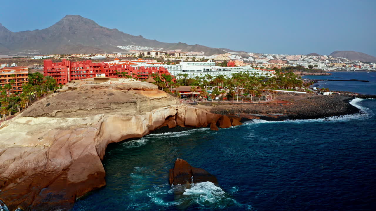 Aerial drone shot over the beautiful volcanic rocky coastline of Tenerife, Canary Islands, Spain. High view of the beach resorts, the turquoise sea and the local towns. Summer holidays destination.