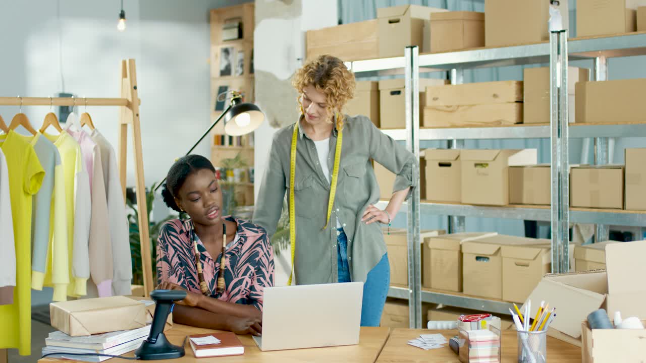 Caucasian and african american designers women speaking while browsing online searching internet on laptop in fashion clothing shop