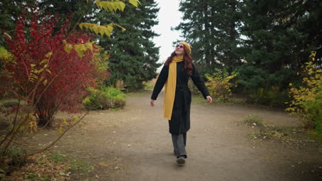 beautiful lady with long red hair wearing black coat, yellow beret, and scarf walking along scenic garden path surrounded by colorful autumn foliage, looking around while placing hand on her head