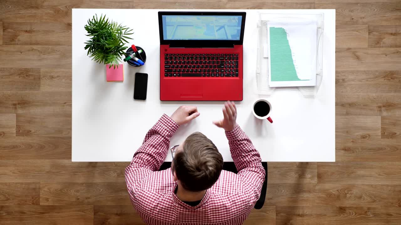 Ung mand med briller taler med nogen via bærbar computer, topshot, siddende bag skrivebord med kaffe og dokumenter