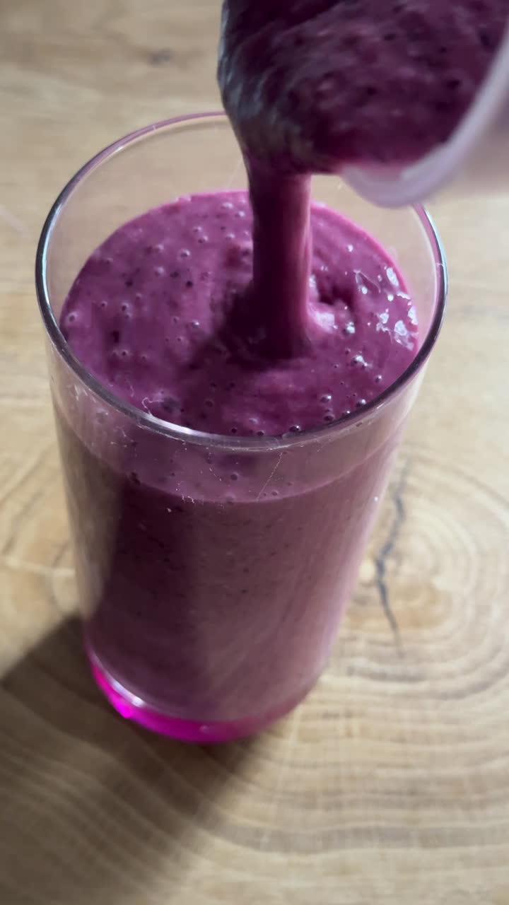 Vertical UGC style shot of thick berry smoothie pouring into a clear glass on wooden table. Natural homemade drink with creamy texture and vibrant fruit color