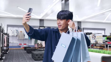 an asian shopping man with shopping bags having video call on mobile phone while standing in clothing store