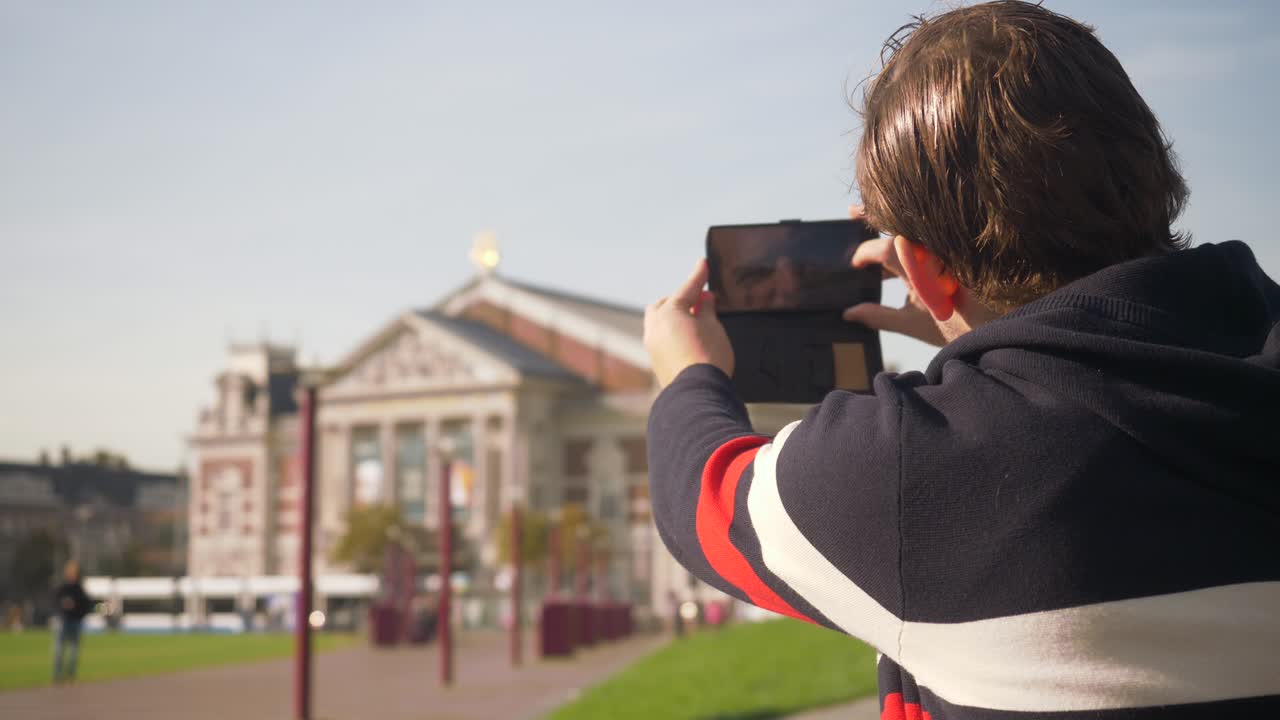 Nice looking young man taking pictures of the Concertgebouw in Amsterdam with the cellphone in a sunny day. Netherlands. Flat plane