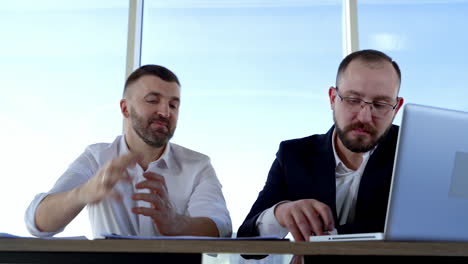 bearded business partners talking in office. male office workers having a conversation about business while sitting at the table by the window.