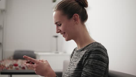 Vista-Más-Cercana-De-Una-Joven-Sonriente-Sentada-En-El-Sofá-De-Casa-Mirando-El-Teléfono-Y-Escribiendo-Un-Mensaje