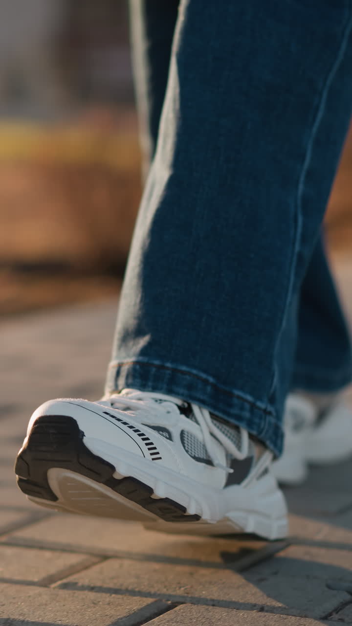 A close-up image of a person wearing jeans and white shoes walking slowly on a paved path in a park. a sense of calmness mixed with sadness, as the deliberate, slow steps reflect a subdued mood
