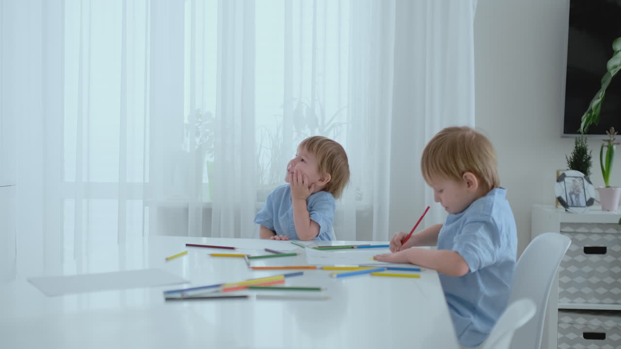 Two boys brothers sit at a table in the living room and draw with pencils on paper. Mom comes up and hugs the boys, smiling and stroking his head. Praises and helps to make the task