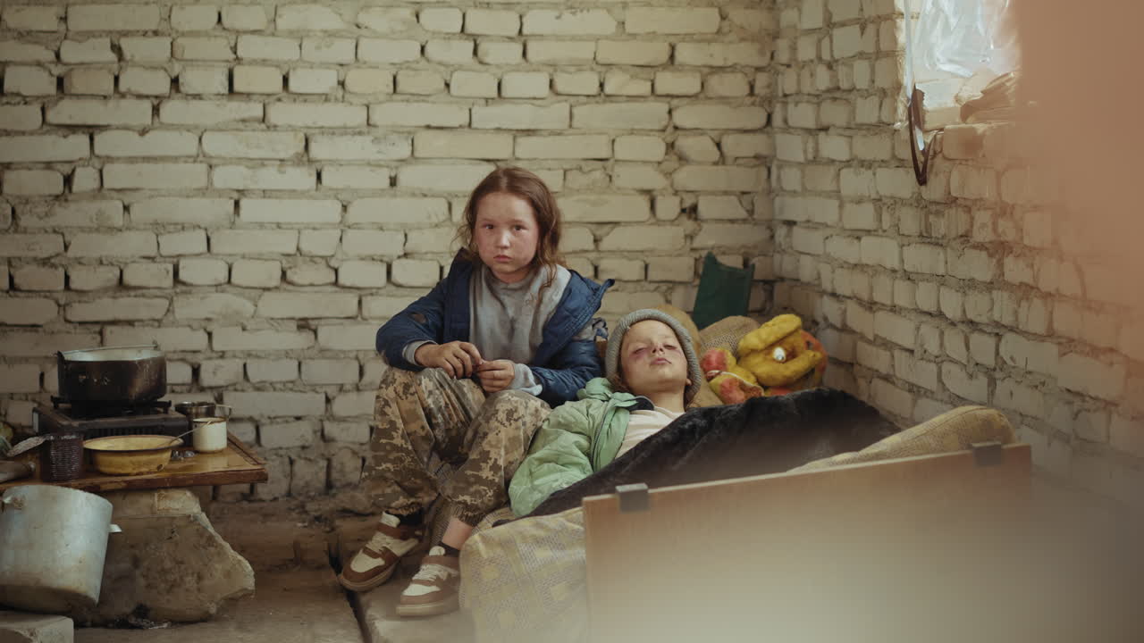 Dirty faced girl in torn clothes sits beside bruised, unwell child on tattered mattress inside neglected brick shelter, nearby scattered cooking pots, old plush toys, and stained dishes