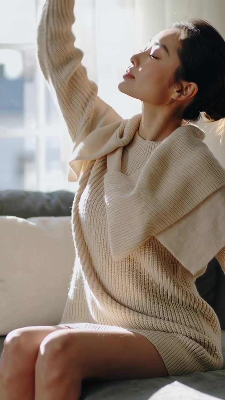 Vertikal video: Sollys falder på kvinde i sweater, der løfter arm ved vinduet, sætter hår, lukker øjnene.