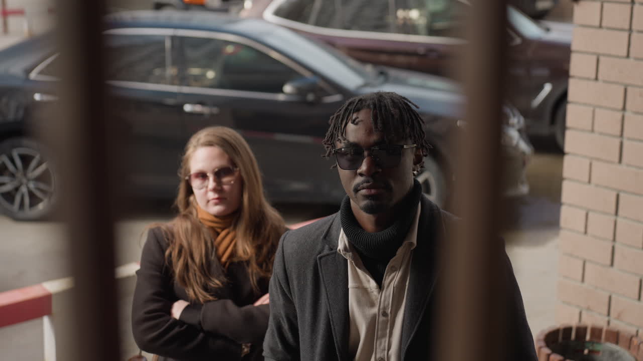 Elegant girl folds arm while stylish man adjusts wool coat behind security grille, both wearing UV glasses at urban entrance near parked cars with business vibe, friends pause before heading inside