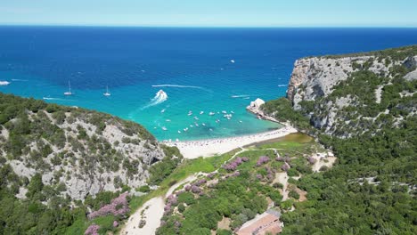 Tourists-and-Boats-at-Cala-Luna-Beach,-Baunei-Coast,-Sardinia,-Italy---Aerial-4k