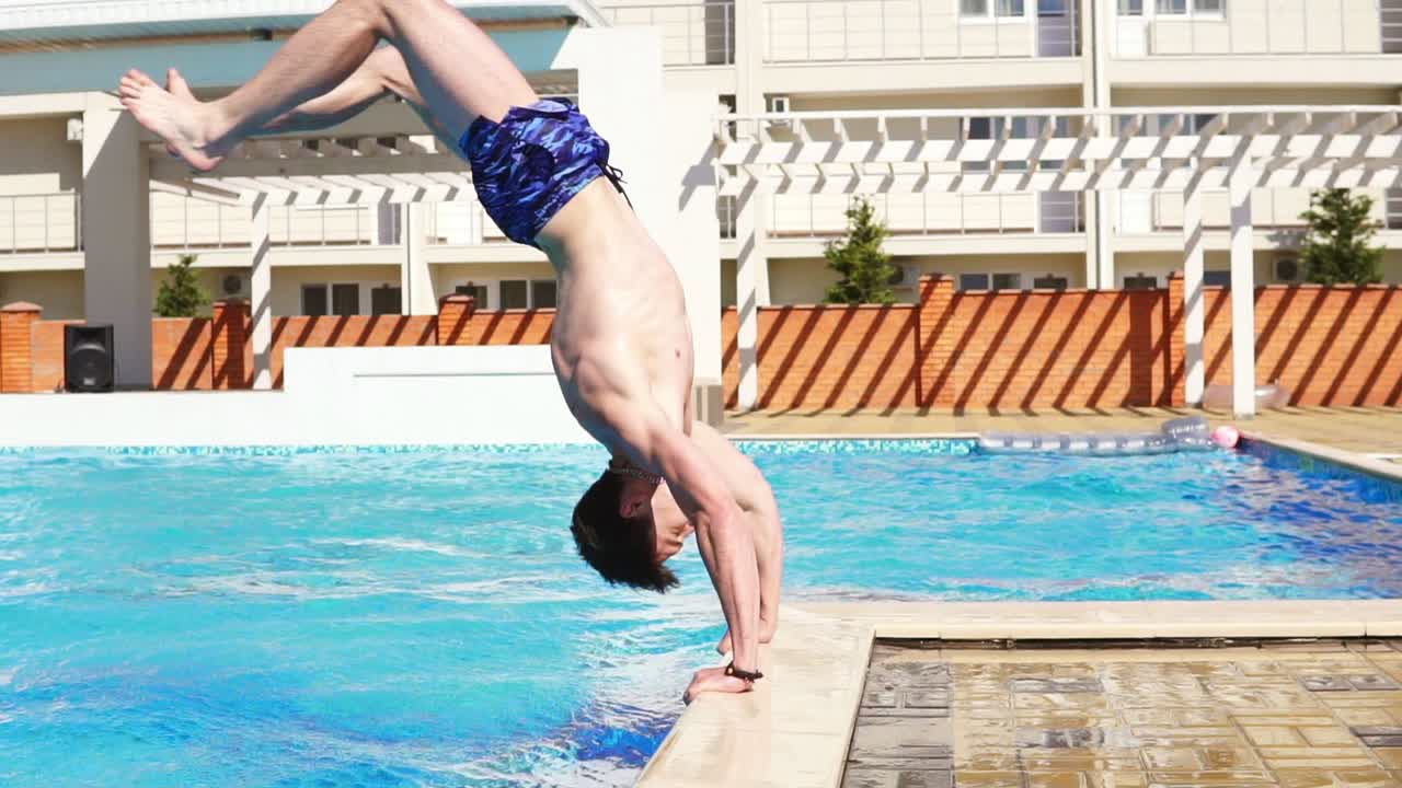 Young athletic man in swim shorts running and jumping to the swimming pool standing on his hands. Slowmotion shot.