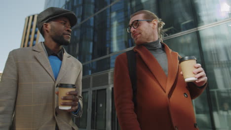 multiethnic male colleagues walking outdoors with coffee and speaking