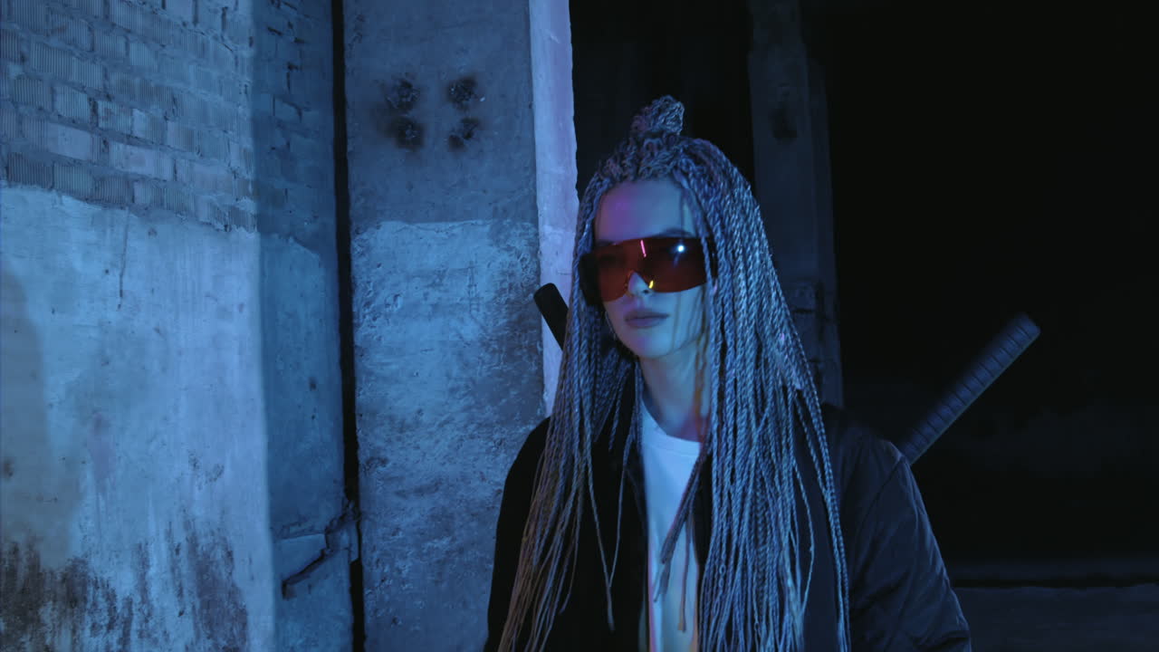 una chica con dreadlocks y katanas en gafas rojas camina a lo largo de una pared de ladrillo de neón