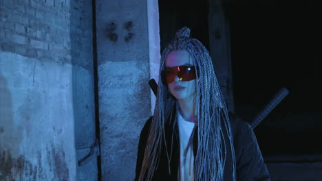 una chica con dreadlocks y katanas en gafas rojas camina a lo largo de una pared de ladrillo de neón