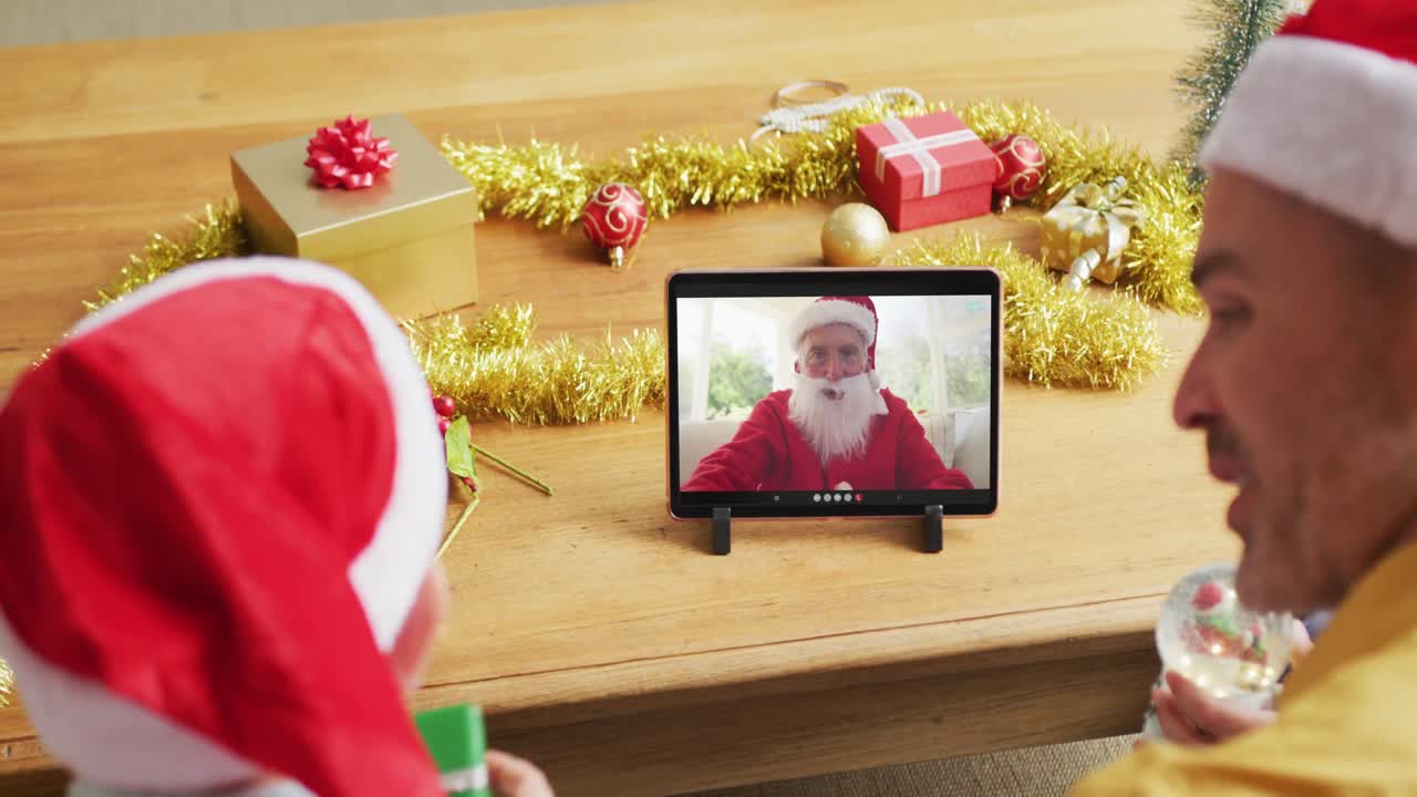 padre y hijo caucásicos con sombreros de santa usando una tableta para una videollamada de navidad con santa en la pantalla