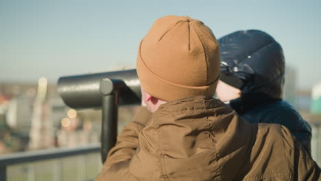 a father holds his son up to a public telescope, guiding him as the looks through it, exploring the distant cityscape, the father ensures a steady view