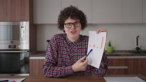 curly - haired with glasses business man sitting at office from home desk looking at camera and pointing at a tablet with financial information displayed in graphical form column graph