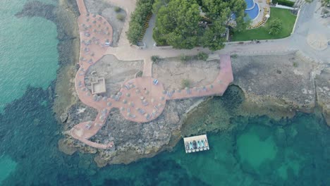 A-coastal-promenade-in-mallorca,-spain,-with-clear-waters-and-scattered-sunbeds,-at-dusk,-aerial-view