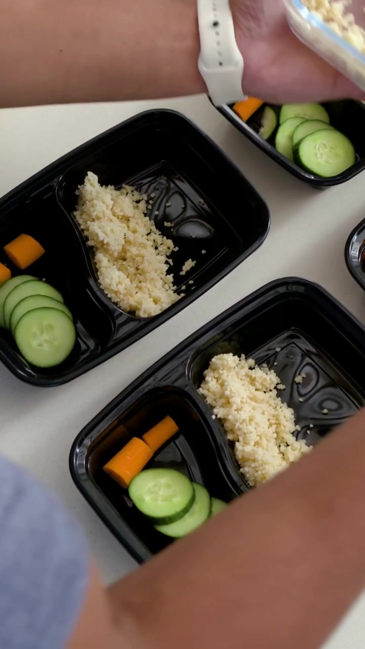 Vertical video: Lifting bowl, cook in blue tee scooping grain into black trays on counter for meals