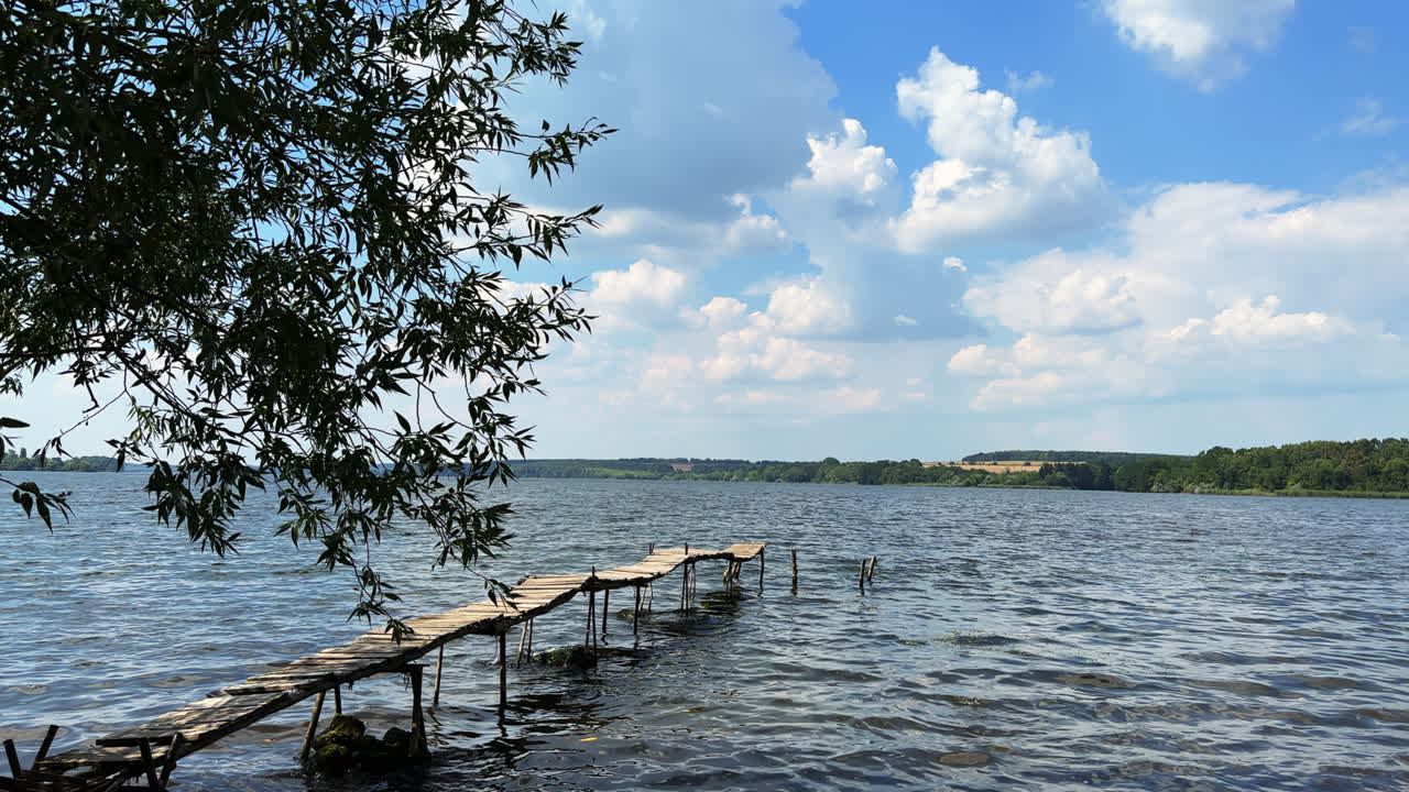 Long wooden old footbridge above the river. Waterscape on sunny day with greenery on the bank at backdrop.