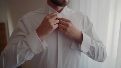 hombre abotonando una camisa blanca, preparándose para una ocasión formal, con la luz del sol filtrando a través de las cortinas