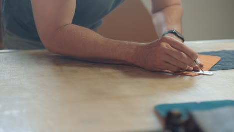 artisan uses metal ruler to draw measurements on a piece of leather that is on his workbench in his workshop