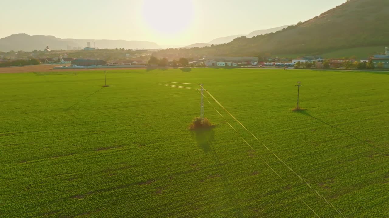 Aerial view of power lines stretching across lush green fields during sunrise. Peaceful rural landscape with the sun illuminating power lines. (4K)