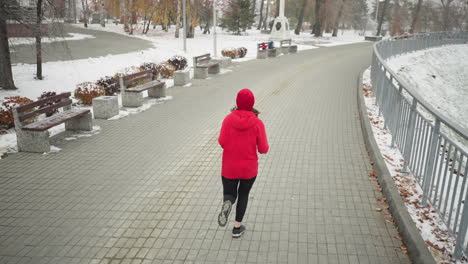 woman jogging along snowy park pathway wearing red hoodie and black leggings, bag placed on distant bench, surrounded by serene winter scenery, trees, bushes, lamp posts, and iron railing