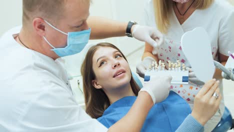 a dentist with an assistant selects dentures by color for the patient. a girl sits in a dental chair and looks in the mirror
