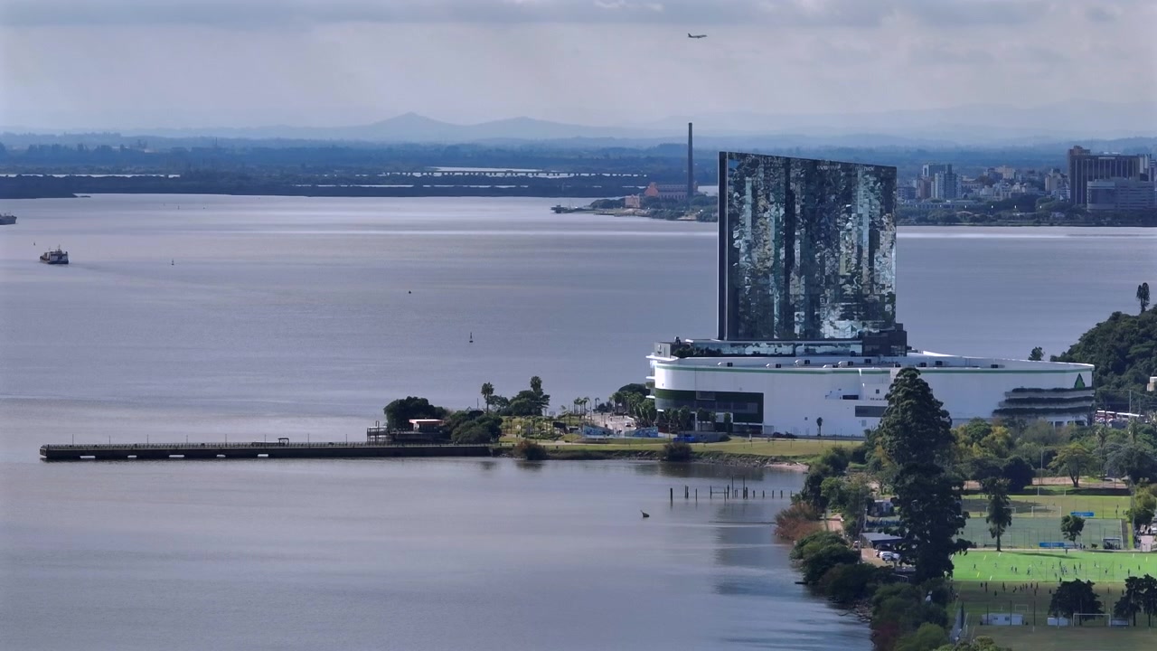 Moderno centro commerciale Pontal con facciata in vetro riflettente sul lago Guaíba a Porto Alegre, con skyline cittadino distante e un aereo che sorvola