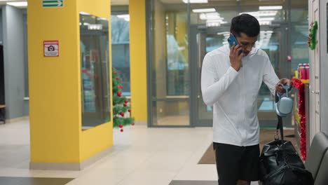 young man in white athletic top and black shorts answers urgent call on blue smartphone, holds headphones, drops leather gym bag and hurries toward exit door in bright modern fitness center lobby