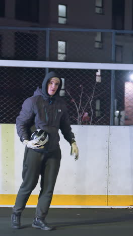 hombre con capucha patea una pelota de fútbol cerca del poste de la portería durante la sesión de entrenamiento nocturna en el campo al aire libre urbano, reflejos de sombras y edificios iluminados de fondo