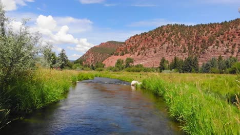 Beautiful-Flowing-River-Lined-With-Green-Grassy-Banks-With-Red-Rocky-Hillside-Cliff