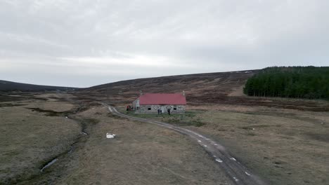 A-drone-flies-backwards-away-from-the-Red-House-Bothy-in-the-Scottish-highlands-as-people-pack-up-bikes-and-talk-outside-the-front-door