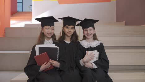 retrato de tres niñas felices con toga y birrete sosteniendo cuadernos y mirando la cámara mientras se sientan en las escaleras en la ceremonia de graduación preescolar