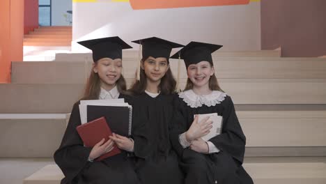 portrait of three happy little girls in cap and gown holding notebooks and looking at the camera while sitting on stairs at the preschool graduation ceremony