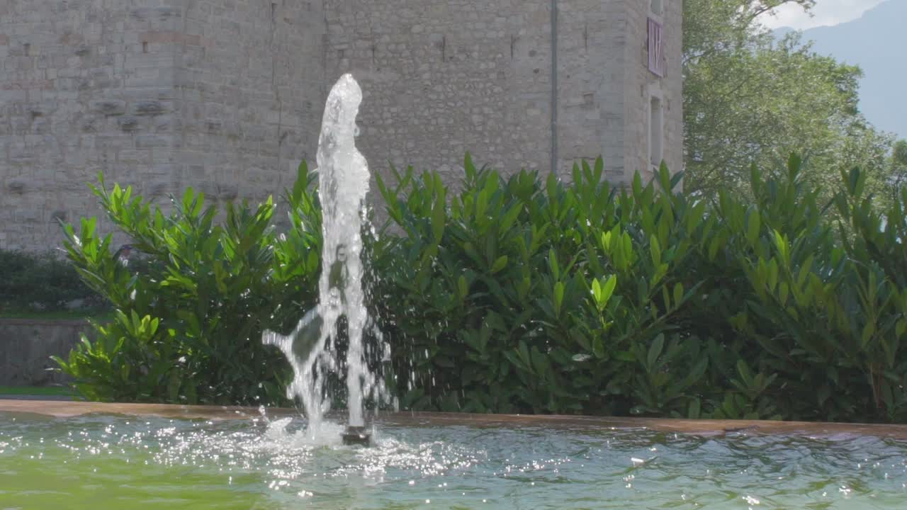 cámara lenta de una pequeña fuente en el centro de riva del garda con algunas plantas verdes y la pared de un antiguo castillo en el fondo