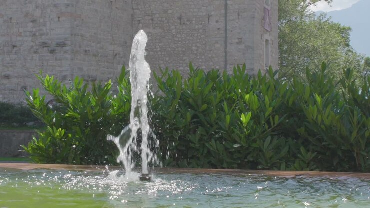 Slowmotion of a small fountain in the center of Riva Del Garda with some green plants and the wall of an old castle in the background