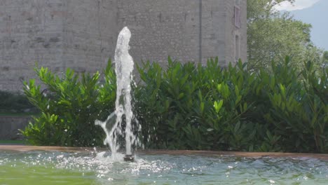 cámara lenta de una pequeña fuente en el centro de riva del garda con algunas plantas verdes y la pared de un antiguo castillo en el fondo