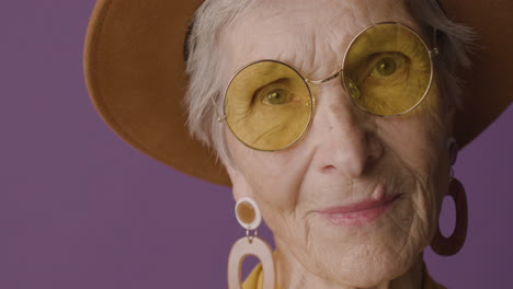 close up view of senior woman with short hair wearing hat, sunglasses and earrings posing and looking at camera on purple background