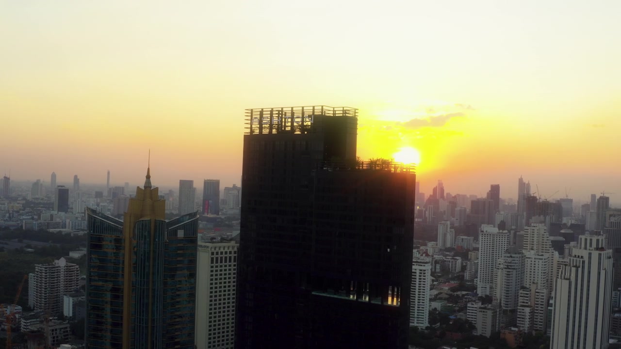 Drone sobrevoa Bangkok enquanto o sol nascente se move por trás de dois edifícios altos, criando uma silhueta urbana quente