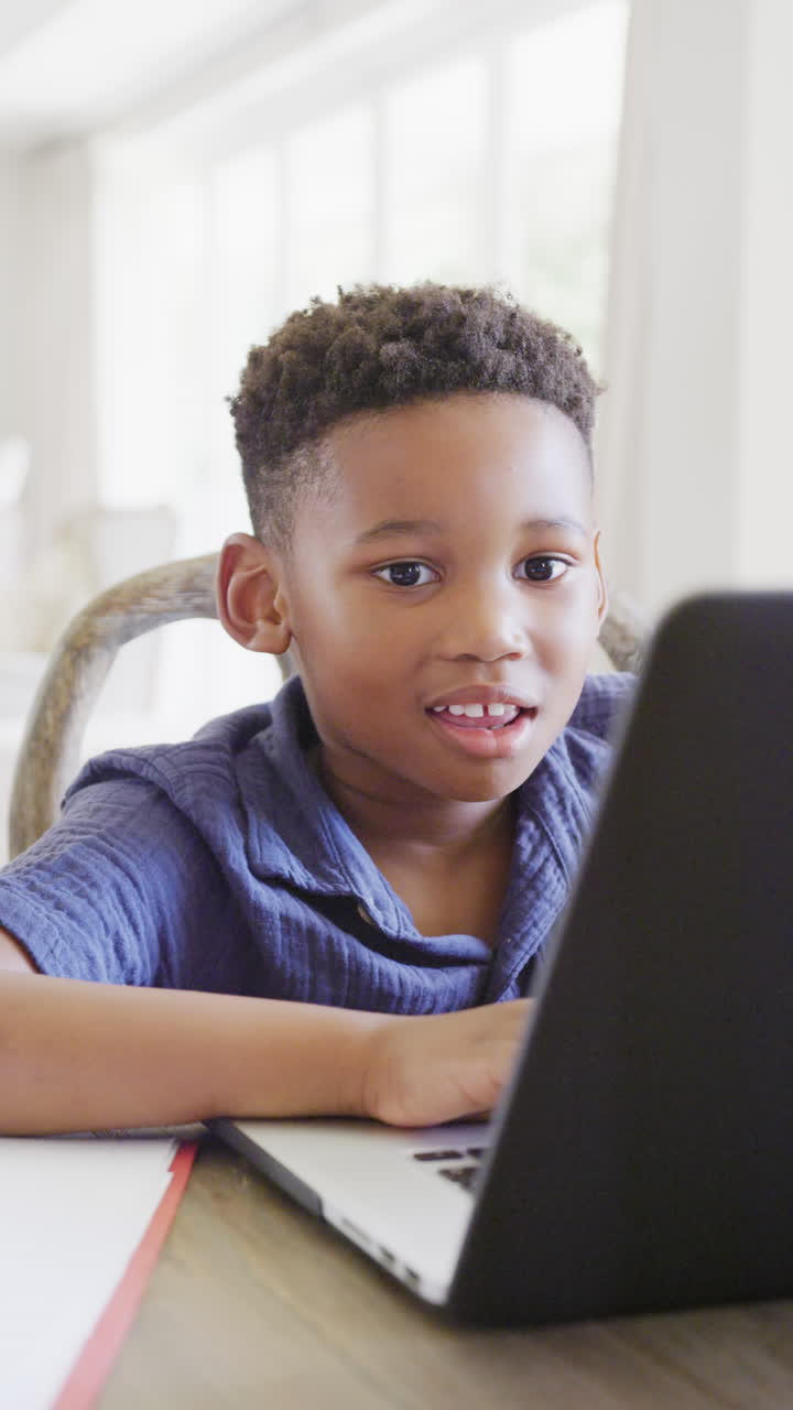Vertical video of happy african american boy using laptop for online lesson, in slow motion