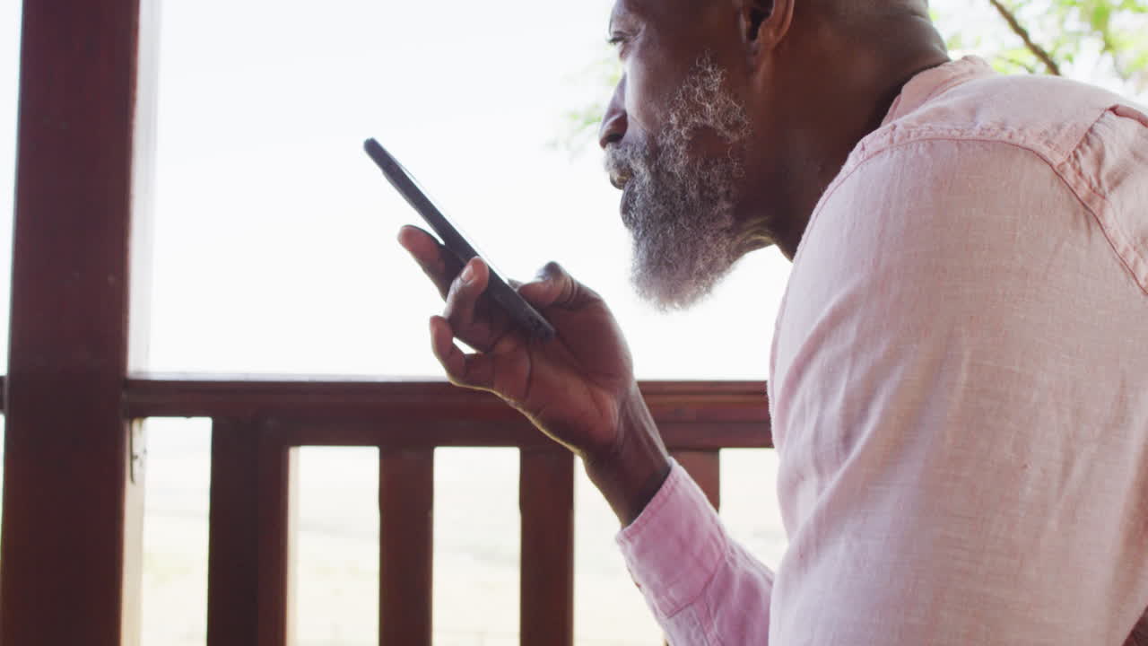 Happy senior african american man in log cabin, talking on smartphone on balcony, slow motion
