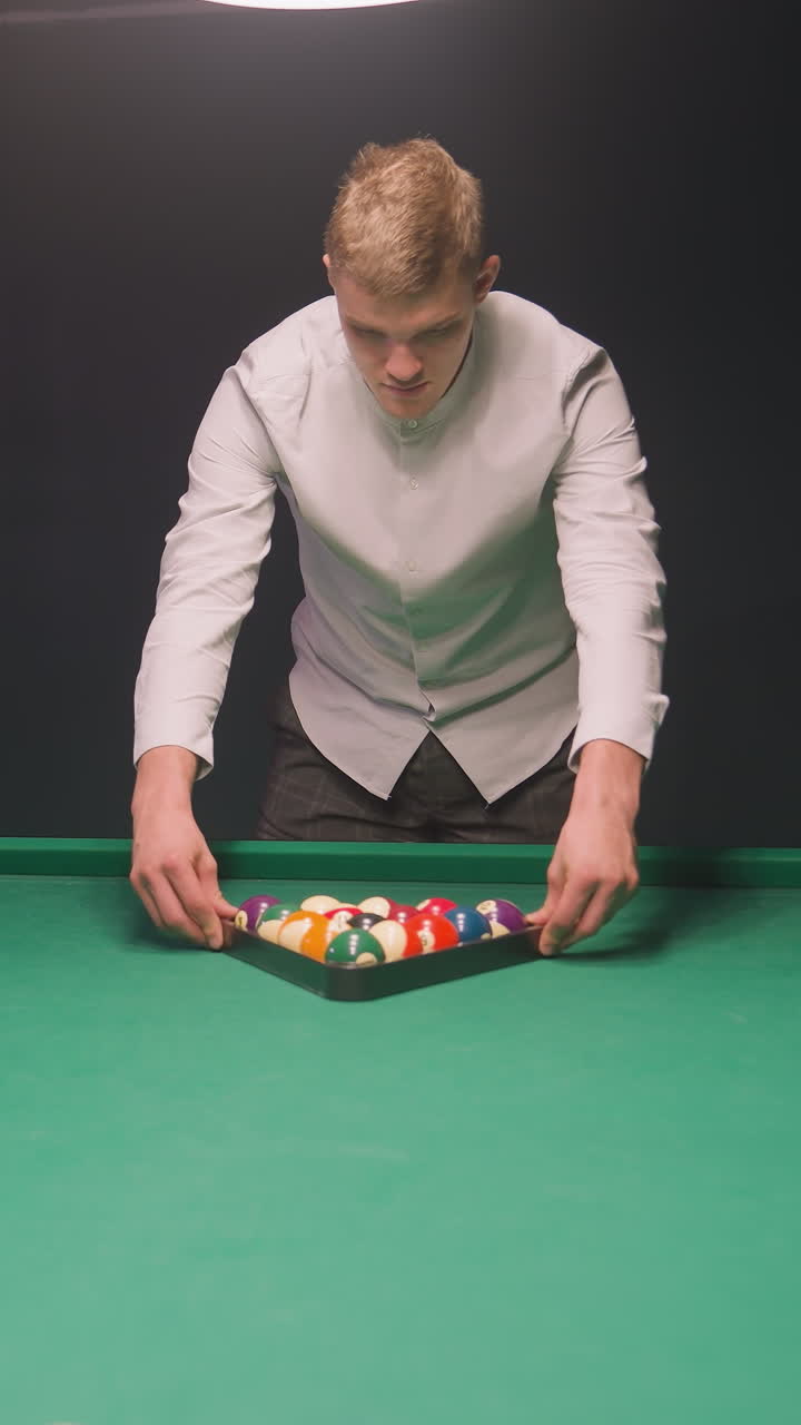 joven rubio con camisa blanca y pantalones gris claro se centra atentamente mientras arregla coloridas bolas de billar en triángulo en una mesa de billar verde vibrante.